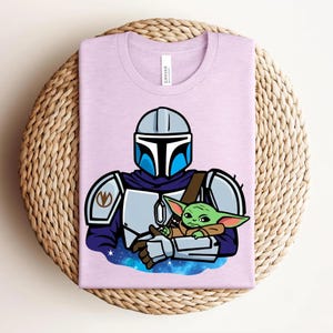 May include: A light purple t-shirt featuring a cartoon illustration of a Mandalorian character holding a green alien. The Mandalorian is wearing a helmet and armor. The alien is small with large ears. The shirt is folded on a woven surface.