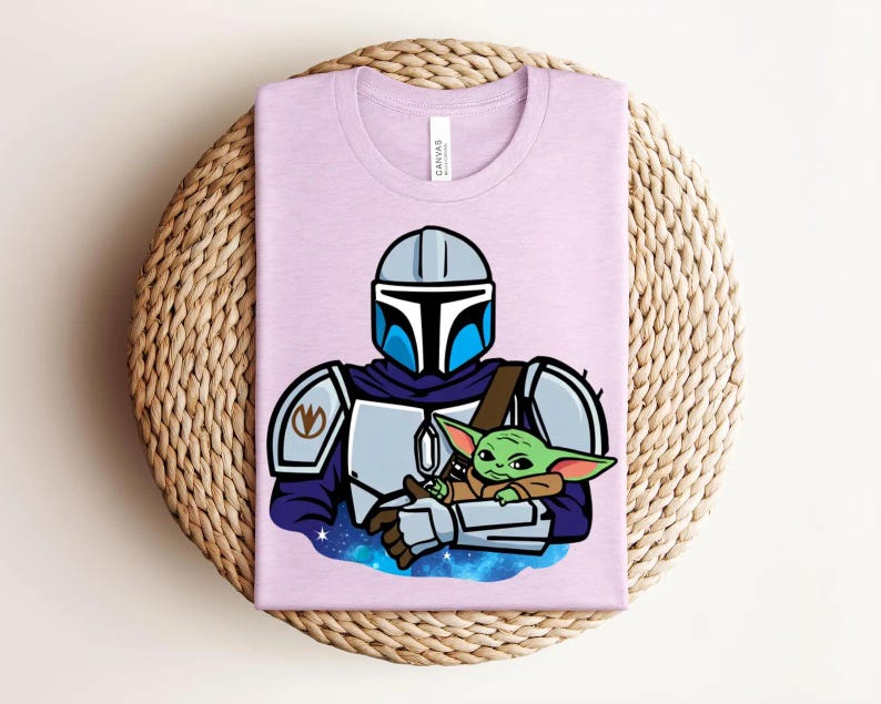 May include: A light purple t-shirt featuring a cartoon illustration of a Mandalorian character holding a green alien. The Mandalorian is wearing a helmet and armor. The alien is small with large ears. The shirt is folded on a woven surface.