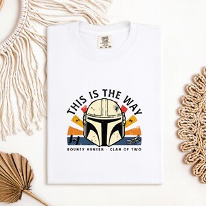 May include: White t-shirt with a Mandalorian helmet graphic and the phrase "THIS IS THE WAY". The design incorporates a sunset-style backdrop and the words "BOUNTY HUNTER" and "CLAN OF TWO".