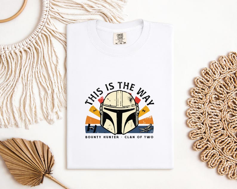 May include: White t-shirt with a Mandalorian helmet graphic and the phrase "THIS IS THE WAY". The design incorporates a sunset-style backdrop and the words "BOUNTY HUNTER" and "CLAN OF TWO".
