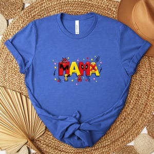 May include: Blue t-shirt with the word "MAMA" in red, yellow, and black, featuring superhero-themed graphics. The shirt has a crew neck and short sleeves. The shirt is displayed on a woven mat with a brown hat in the background.