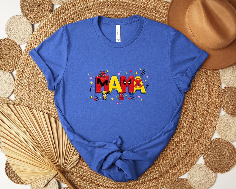 May include: Blue t-shirt with the word "MAMA" in red, yellow, and black, featuring superhero-themed graphics. The shirt has a crew neck and short sleeves. The shirt is displayed on a woven mat with a brown hat in the background.