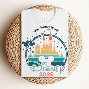 Op de afbeelding: Wit T-shirt met een kleurrijke afbeelding van een kasteel, Mickey Mouse oren en de tekst "Walt Disney World" en "Disney 2026". Het ontwerp heeft een regenboog aan kleuren en sterren, met een blauwe omtrek.