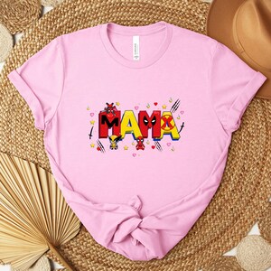 May include: A light pink t-shirt with the word "MAMA" in colorful, comic-style lettering. The design includes cartoon characters and symbols, with hearts and stars scattered around the text. The shirt is folded.