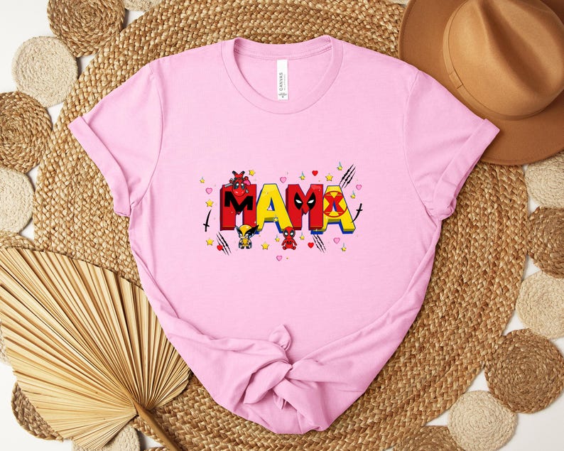 May include: A light pink t-shirt with the word "MAMA" in colorful, comic-style lettering. The design includes cartoon characters and symbols, with hearts and stars scattered around the text. The shirt is folded.