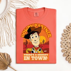 K&ouml;nnte beinhalten: Korallenfarbenes T-Shirt mit einer Cartoon-Illustration eines Cowboys und dem Text "HOWDY PARTNER" und "THERE'S A SHERIFF IN TOWN". Das Design beinhaltet eine Sonnenuntergangsszene mit Kakteen und einer Sonne. Das T-Shirt hat einen Rundhalsausschnitt.