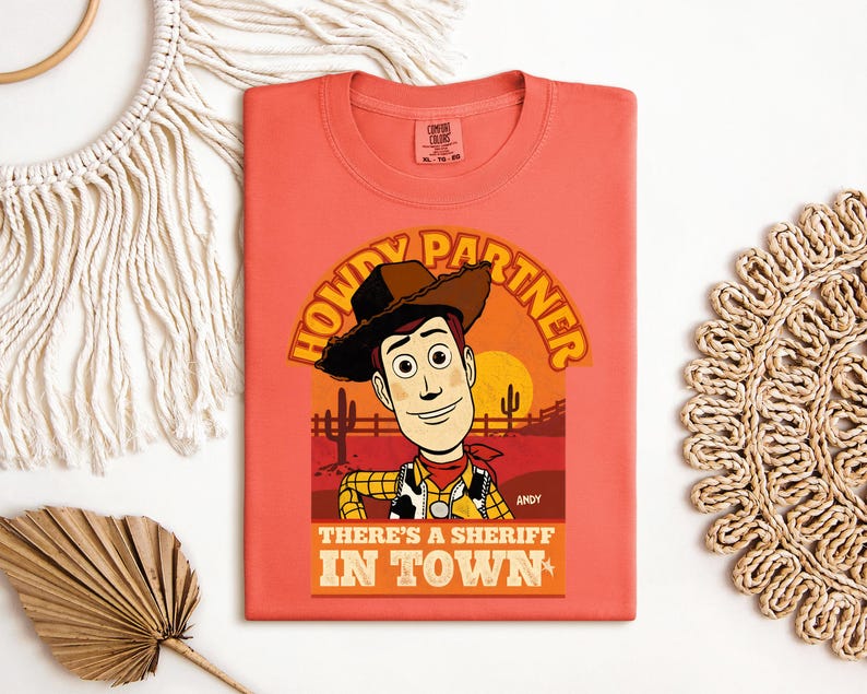 K&ouml;nnte beinhalten: Korallenfarbenes T-Shirt mit einer Cartoon-Illustration eines Cowboys und dem Text "HOWDY PARTNER" und "THERE'S A SHERIFF IN TOWN". Das Design beinhaltet eine Sonnenuntergangsszene mit Kakteen und einer Sonne. Das T-Shirt hat einen Rundhalsausschnitt.