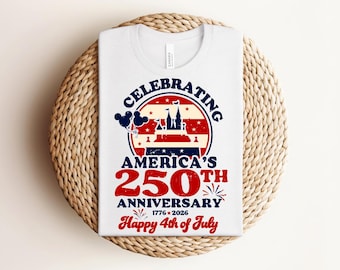 May include: Light blue t-shirt with a graphic celebrating America's 250th Anniversary. The design includes a castle, balloons, and the text "Happy 4th of July" in red and white. The shirt is laid on a woven, circular mat.