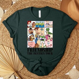 May include: Dark green t-shirt featuring a colorful graphic of various cartoon characters in a grid pattern. The text "FRIENDSHIP THE BESTIES TOUR" is printed below the graphic. The shirt is displayed on a woven surface.