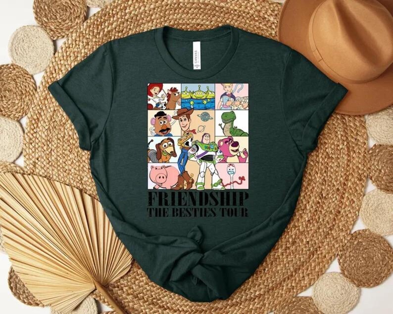 May include: Dark green t-shirt featuring a colorful graphic of various cartoon characters in a grid pattern. The text "FRIENDSHIP THE BESTIES TOUR" is printed below the graphic. The shirt is displayed on a woven surface.