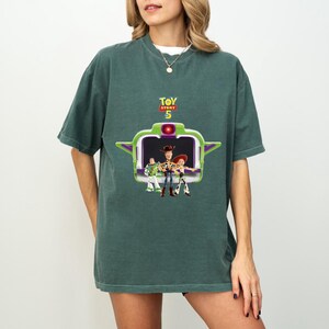 K&ouml;nnte beinhalten: Gr&uuml;nes T-Shirt mit einem Grafikmotiv aus dem Film Toy Story 5. Die Grafik zeigt Buzz Lightyear, Woody und Jessie in einem gr&uuml;nen und lila Rahmen. Die Worte "TOY STORY 5" stehen in Gelb und Rot &uuml;ber der Grafik.
