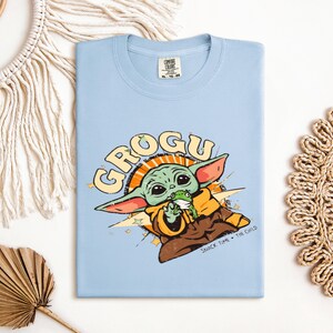 May include: A light blue t-shirt featuring a graphic of Grogu, also known as The Child, from The Mandalorian. The design includes the word "GROGU" in a retro font, and Grogu is depicted holding a frog. The text "SNACK TIME - THE CHILD" is also included.