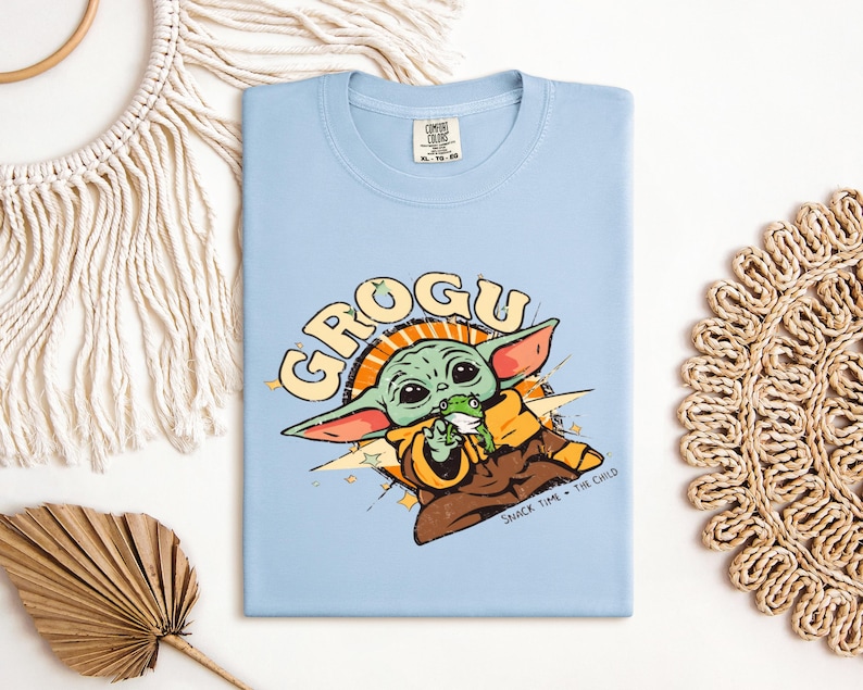 May include: A light blue t-shirt featuring a graphic of Grogu, also known as The Child, from The Mandalorian. The design includes the word "GROGU" in a retro font, and Grogu is depicted holding a frog. The text "SNACK TIME - THE CHILD" is also included.
