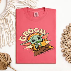 May include: A coral-colored t-shirt featuring a graphic of Grogu, also known as The Child, from The Mandalorian. The design includes the word "GROGU" in a retro font, and an illustration of Grogu holding a frog. The shirt has a crew neck.