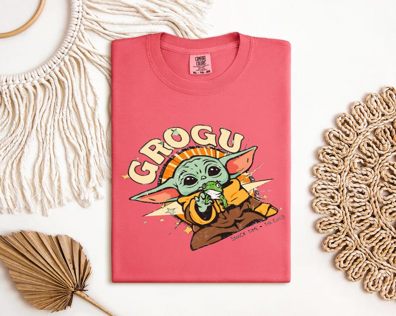 May include: A coral-colored t-shirt featuring a graphic of Grogu, also known as The Child, from The Mandalorian. The design includes the word "GROGU" in a retro font, and an illustration of Grogu holding a frog. The shirt has a crew neck.