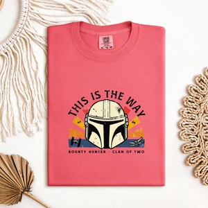 May include: A coral t-shirt with a graphic of a Mandalorian helmet and the phrase "THIS IS THE WAY." The design includes the text "BOUNTY HUNTER - CLAN OF TWO" below the helmet. The graphic has yellow, orange, and blue accents.
