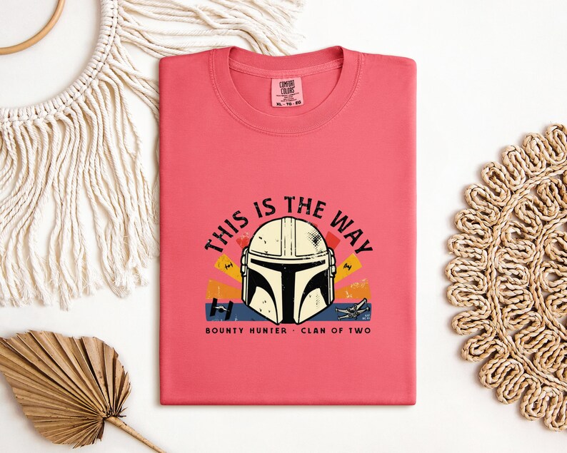 May include: A coral t-shirt with a graphic of a Mandalorian helmet and the phrase "THIS IS THE WAY." The design includes the text "BOUNTY HUNTER - CLAN OF TWO" below the helmet. The graphic has yellow, orange, and blue accents.