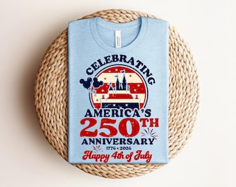 May include: Pale blue t-shirt featuring a graphic commemorating America's 250th Anniversary. The design includes a castle, balloons, and the text "Happy 4th of July" in red and white. The shirt is placed on a woven, circular mat.