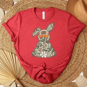 May include: A red t-shirt featuring a cartoon bunny with a camouflage pattern and a duck-billed cap. The bunny is sitting, and the shirt is styled on a woven mat with a hat.