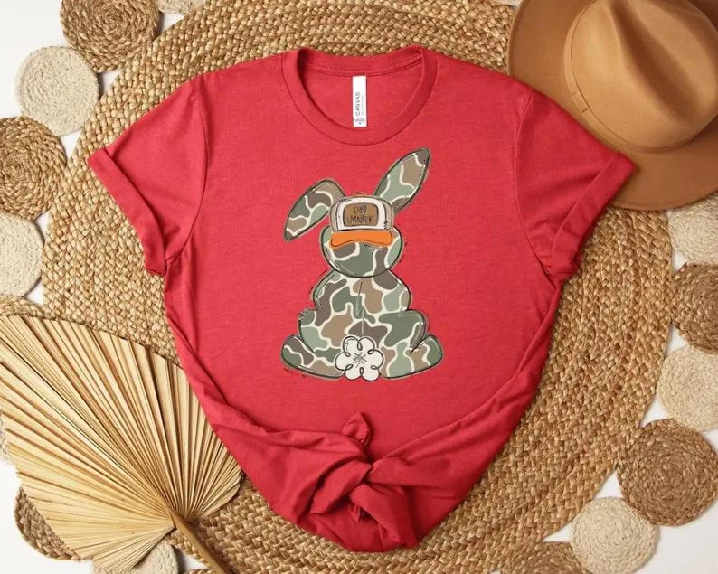 May include: A red t-shirt featuring a cartoon bunny with a camouflage pattern and a duck-billed cap. The bunny is sitting, and the shirt is styled on a woven mat with a hat.