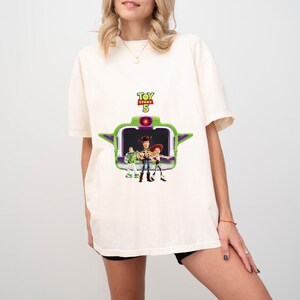 K&ouml;nnte beinhalten: Cremefarbenes T-Shirt mit einer Grafik aus dem Film Toy Story 5. Die Grafik zeigt Woody, Buzz Lightyear und Jessie vor einer gr&uuml;n-violetten raumschiffartigen Struktur. Der Text "Toy Story 5" befindet sich &uuml;ber der Grafik.