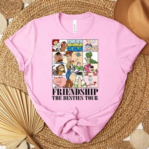 May include: A light pink t-shirt featuring a graphic of various cartoon characters in a grid, with the text "FRIENDSHIP THE BESTIES TOUR" in black. The shirt is made of soft material.