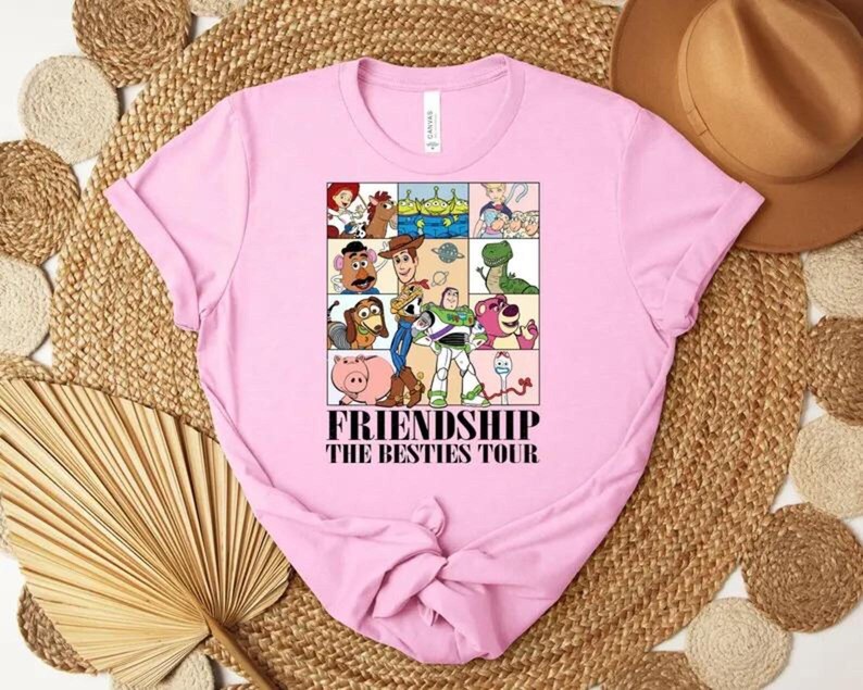 May include: A light pink t-shirt featuring a graphic of various cartoon characters in a grid, with the text "FRIENDSHIP THE BESTIES TOUR" in black. The shirt is made of soft material.