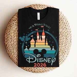 Op de afbeelding: Zwart T-shirt met een kleurrijk kasteelontwerp, de tekst "Disney World" en "Disney 2026". Het ontwerp bevat een Mickey Mouse silhouet en een regenboogkleurig kasteel. Het shirt ligt op een geweven, cirkelvormig oppervlak.