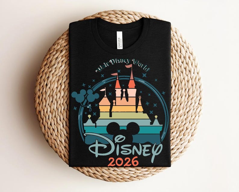Op de afbeelding: Zwart T-shirt met een kleurrijk kasteelontwerp, de tekst "Disney World" en "Disney 2026". Het ontwerp bevat een Mickey Mouse silhouet en een regenboogkleurig kasteel. Het shirt ligt op een geweven, cirkelvormig oppervlak.