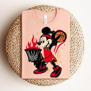 May include: A light peach-colored t-shirt featuring a cartoon Mickey Mouse playing basketball. Mickey is wearing a red uniform and headband, with a basketball in hand. The design includes a flaming basketball hoop and star accents.