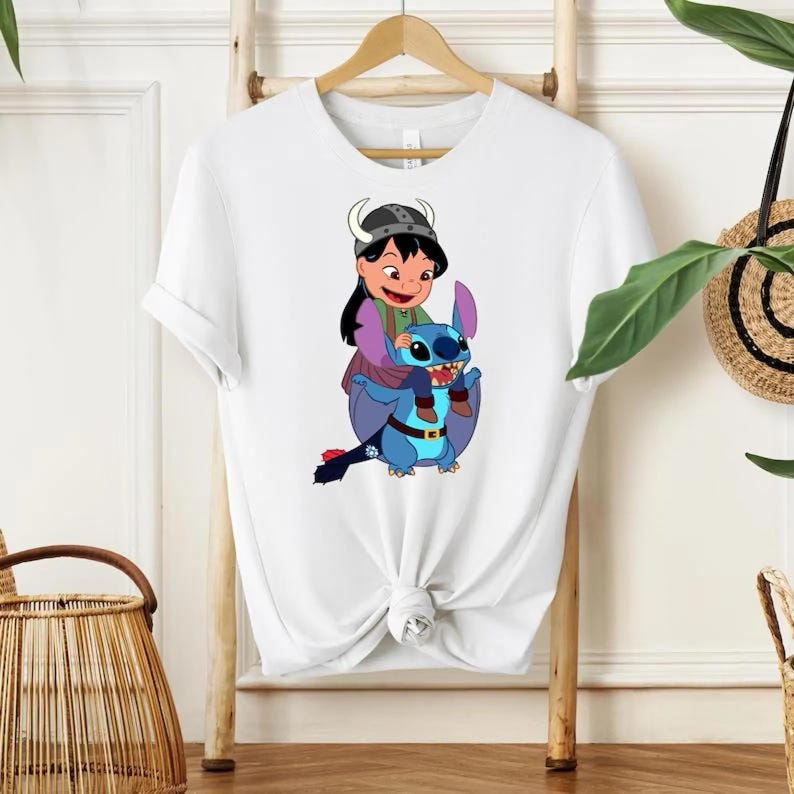 Może przedstawiać: Biała koszulka z kolorową grafiką przedstawiającą dziewczynkę w hełmie wikinga jadącą na niebieskim, obcym stworzeniu. Grafika jest wyśrodkowana na koszulce, kt&oacute;ra wisi na drewnianym wieszaku.