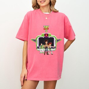K&ouml;nnte beinhalten: Ein pinkfarbenes T-Shirt mit einer Grafik aus dem Film Toy Story 5. Die Grafik zeigt Buzz Lightyear, Woody und Jessie vor einer gr&uuml;n-wei&szlig;en raumschiffartigen Struktur. Die Worte "Toy Story 5" stehen &uuml;ber der Grafik.