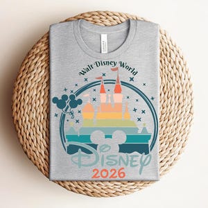 Op de afbeelding: Een grijs t-shirt met een kleurrijke afbeelding van het kasteel van Assepoester, de tekst "Walt Disney World" en "Disney 2026". Het ontwerp bevat Mickey Mouse oren en ballonnen, alles in een cirkelvormig kader.