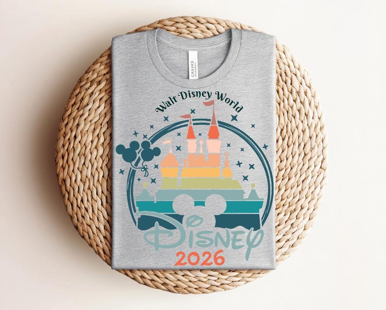 Op de afbeelding: Een grijs t-shirt met een kleurrijke afbeelding van het kasteel van Assepoester, de tekst "Walt Disney World" en "Disney 2026". Het ontwerp bevat Mickey Mouse oren en ballonnen, alles in een cirkelvormig kader.