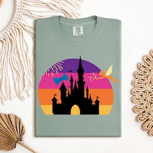 May include: Sage green t-shirt featuring a colorful graphic of a castle silhouette against a sunset backdrop. A fairy and fireworks are also included in the design. The shirt has a crew neck and a tag in the upper center.