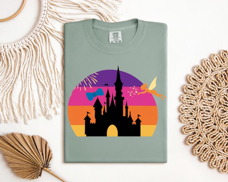 May include: Sage green t-shirt featuring a colorful graphic of a castle silhouette against a sunset backdrop. A fairy and fireworks are also included in the design. The shirt has a crew neck and a tag in the upper center.