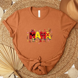 May include: A rust-colored t-shirt featuring the word "MAMA" in a colorful, comic-book style font. The design incorporates superhero-themed elements, including characters and symbols. The shirt is made of soft material and is suitable for casual wear.