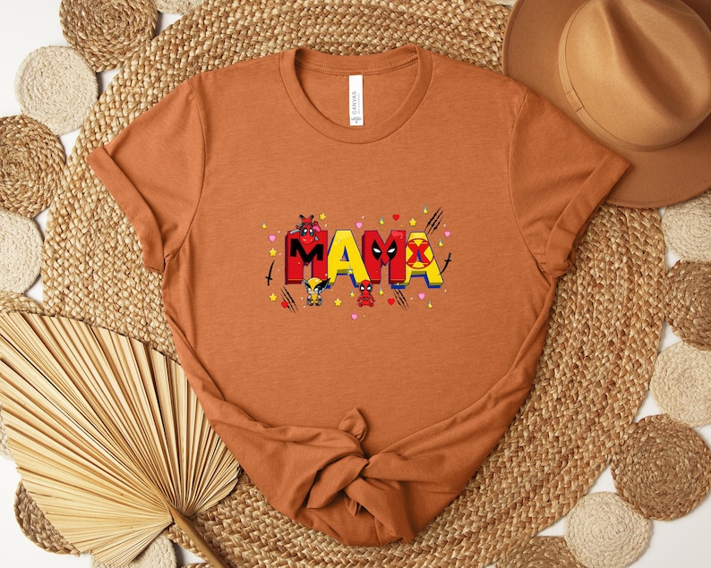 May include: A rust-colored t-shirt featuring the word "MAMA" in a colorful, comic-book style font. The design incorporates superhero-themed elements, including characters and symbols. The shirt is made of soft material and is suitable for casual wear.