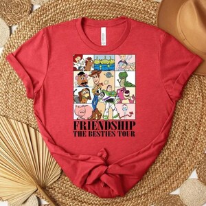 May include: Red t-shirt featuring a colorful graphic of cartoon characters in a grid, with the words "FRIENDSHIP THE BESTIES TOUR" in a bold font. The shirt is made of a soft material and has a classic crew neck design.