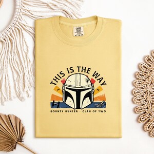 May include: Mustard yellow t-shirt with a graphic of a Mandalorian helmet and the phrase "THIS IS THE WAY". The design incorporates a sunset and the words "BOUNTY HUNTER" and "CLAN OF TWO".