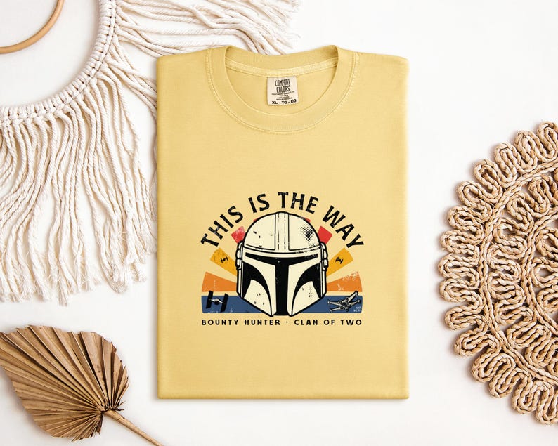 May include: Mustard yellow t-shirt with a graphic of a Mandalorian helmet and the phrase "THIS IS THE WAY". The design incorporates a sunset and the words "BOUNTY HUNTER" and "CLAN OF TWO".