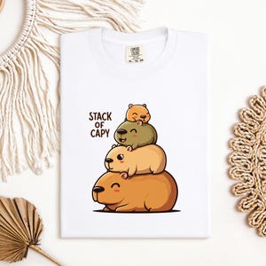 May include: White t-shirt featuring a cartoon illustration of three capybaras stacked on top of each other. The text "STACK OF CAPY" is printed above the capybaras. The shirt has a crew neck and a tag on the upper right.