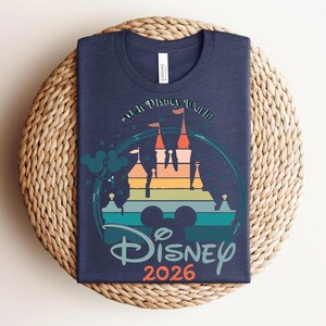 Op de afbeelding: Marineblauw t-shirt met een kleurrijke afbeelding van een kasteel, de tekst "Walt Disney World" en "Disney 2026". Het ontwerp bevat Mickey Mouse oren en een regenboogkleurig kasteelsilhouet.