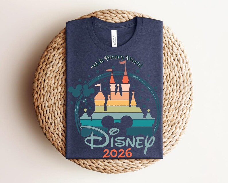 Op de afbeelding: Marineblauw t-shirt met een kleurrijke afbeelding van een kasteel, de tekst "Walt Disney World" en "Disney 2026". Het ontwerp bevat Mickey Mouse oren en een regenboogkleurig kasteelsilhouet.