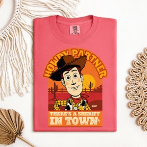 K&ouml;nnte beinhalten: Korallenfarbenes T-Shirt mit einem Cartoon-Cowboy-Design. Der Aufdruck enth&auml;lt den Text "HOWDY PARTNER" und "THERE'S A SHERIFF IN TOWN". Der Cowboy tr&auml;gt einen Hut, eine Weste und ein rotes Halstuch. Der Hintergrund zeigt eine W&uuml;stenlandschaft mit Sonnenuntergang.