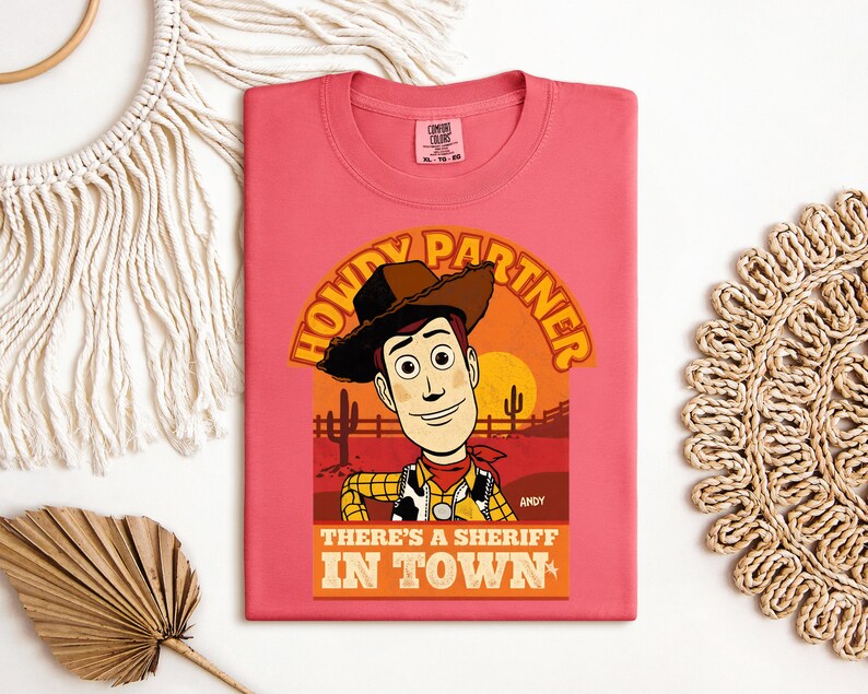 K&ouml;nnte beinhalten: Korallenfarbenes T-Shirt mit einem Cartoon-Cowboy-Design. Der Aufdruck enth&auml;lt den Text "HOWDY PARTNER" und "THERE'S A SHERIFF IN TOWN". Der Cowboy tr&auml;gt einen Hut, eine Weste und ein rotes Halstuch. Der Hintergrund zeigt eine W&uuml;stenlandschaft mit Sonnenuntergang.