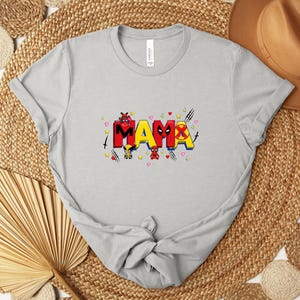 May include: A light gray t-shirt with the word "MAMA" in a colorful, comic book style. The letters are red, yellow, and black, with superhero-themed graphics. The shirt is made of soft material.