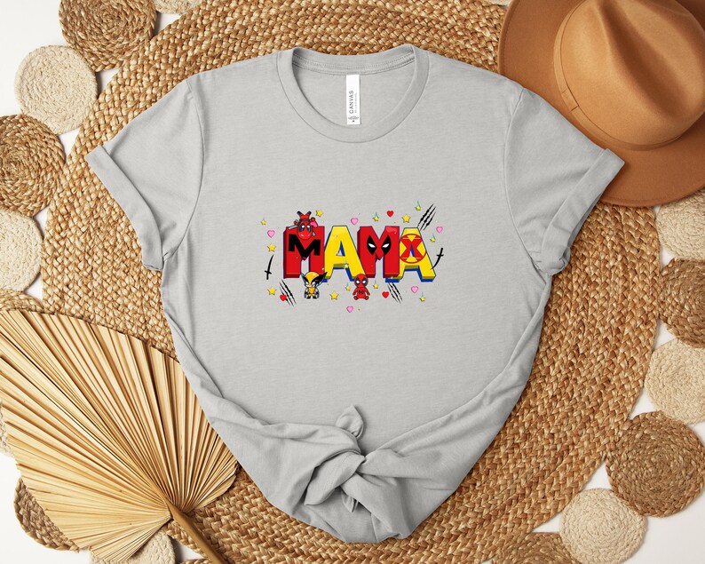 May include: A light gray t-shirt with the word "MAMA" in a colorful, comic book style. The letters are red, yellow, and black, with superhero-themed graphics. The shirt is made of soft material.