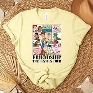 May include: A light yellow t-shirt featuring a colorful graphic of cartoon characters in a grid pattern. The graphic includes characters from the Toy Story franchise. The text "FRIENDSHIP THE BESTIES TOUR" is printed below the graphic.