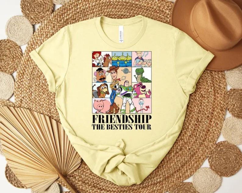 May include: A light yellow t-shirt featuring a colorful graphic of cartoon characters in a grid pattern. The graphic includes characters from the Toy Story franchise. The text "FRIENDSHIP THE BESTIES TOUR" is printed below the graphic.